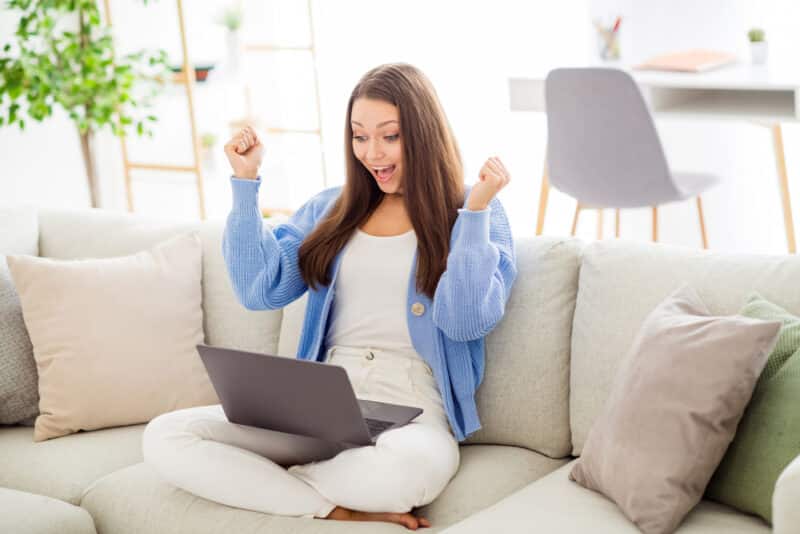 Portrait of pretty cheerful girl sitting on divan using laptop having fun rejoicing start-up in light flat house apartment indoor